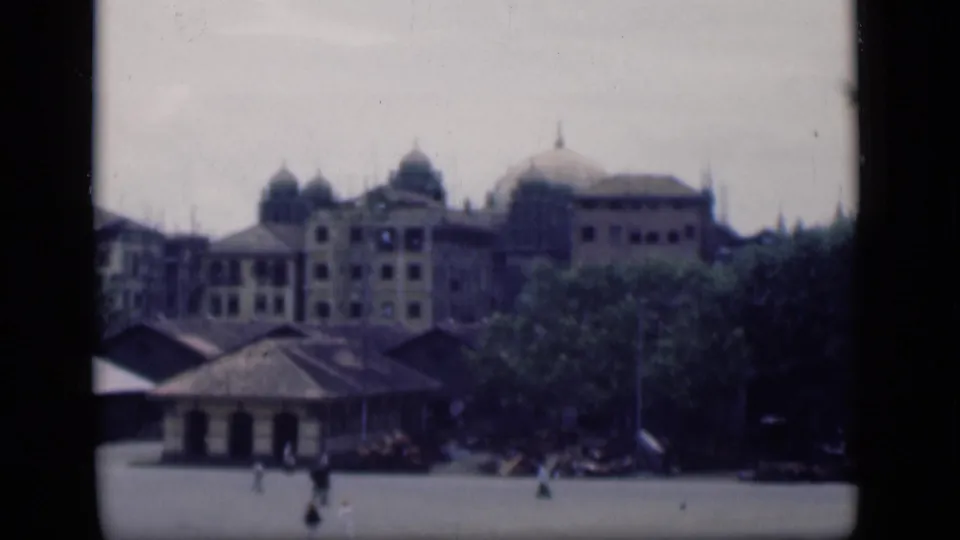 1946 Bombay home-movie view of a domed colonial-style building beyond trees and low structures, with small pedestrians in the foreground.