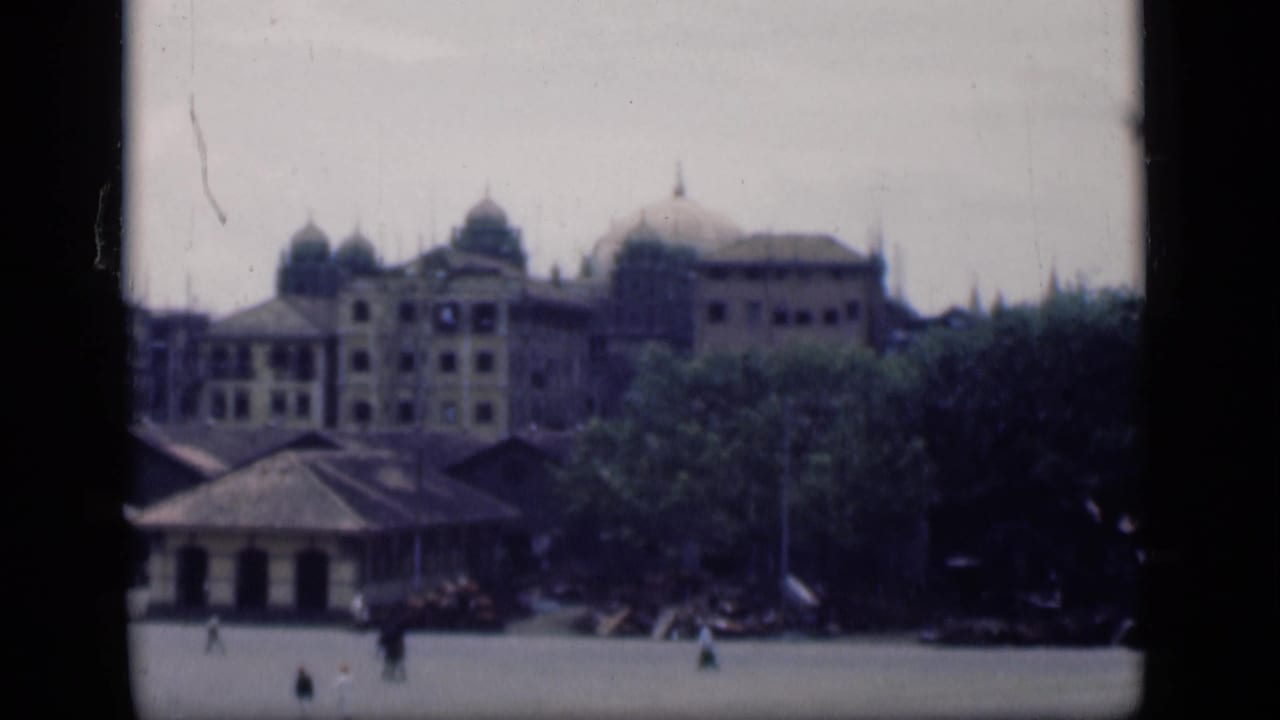Distant 1946 Bombay street view showing colonial-style domes, trees, low foreground roofs, and small figures moving across open pavement.