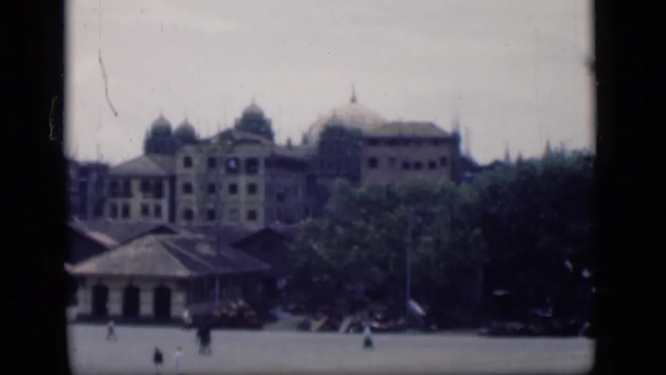Distant 1946 Bombay street view showing colonial-style domes, trees, low foreground roofs, and small figures moving across open pavement.