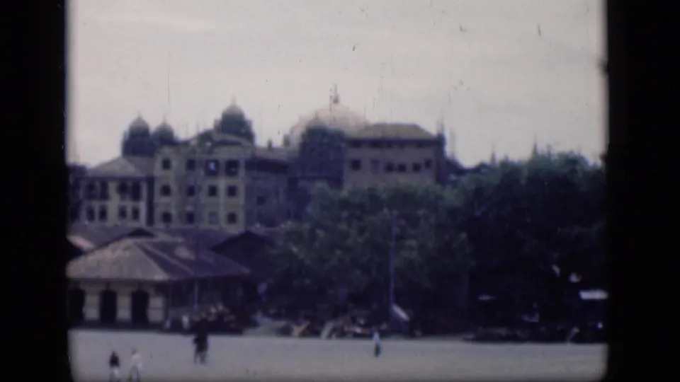 1946 Bombay 8mm frame with domed architecture behind trees and a quiet open area where a few pedestrians are visible.