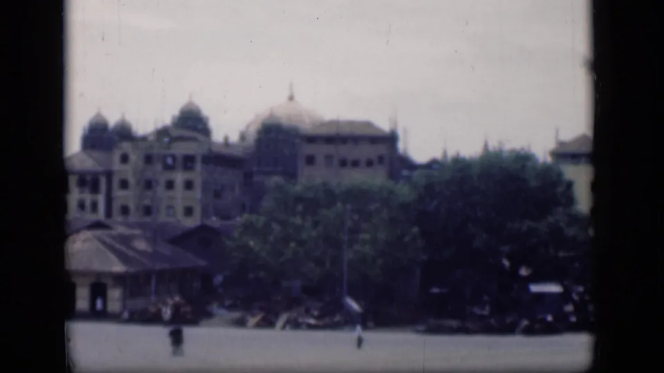 Distant 1946 Bombay view of domed colonial-style architecture, dense trees, low structures, and pedestrians crossing the open foreground.