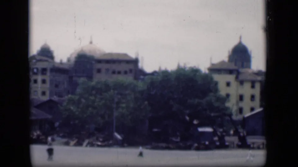 1946 Bombay skyline view with domed rooftops, pale facades, foreground trees, and small indistinct people in the lower frame.