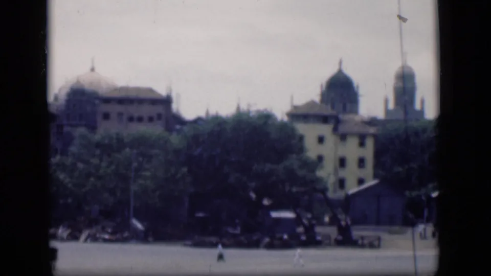 1946 Bombay cityscape showing domed rooftops, tree cover, pale buildings, and indistinct people crossing the lower frame.