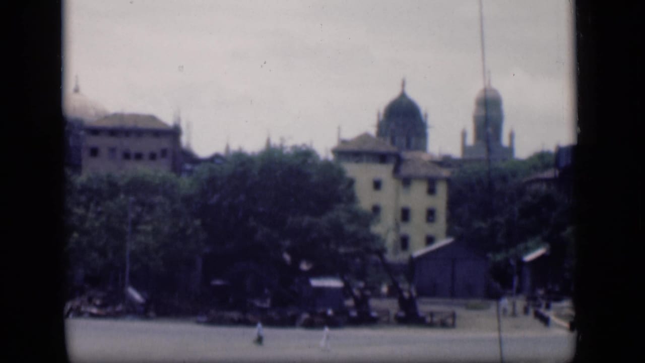 1946 Bombay cityscape with domed buildings, trees, and a wide paved plaza seen from a distant home-movie viewpoint.