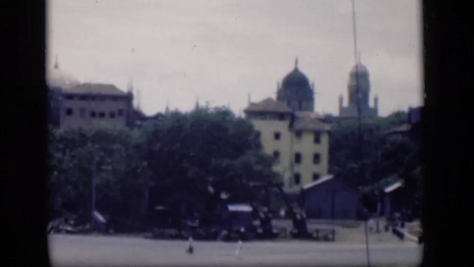 1946 Bombay cityscape with domed buildings, trees, and a wide paved plaza seen from a distant home-movie viewpoint.