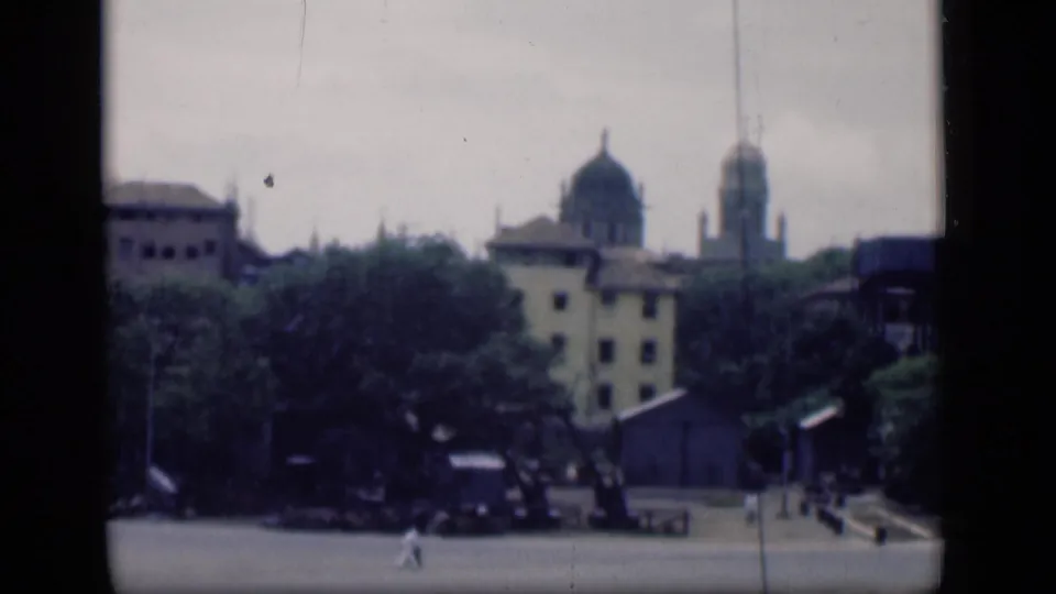 Distant 1946 Bombay plaza view showing domed architecture above trees and scattered pedestrians in the foreground.