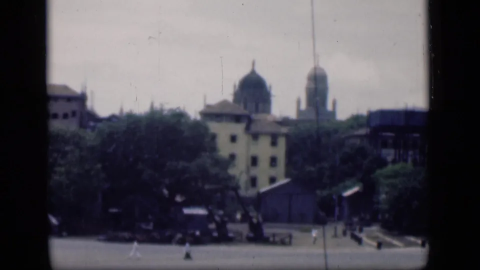1946 Bombay exterior with domed buildings behind trees and small figures moving across a broad paved foreground.