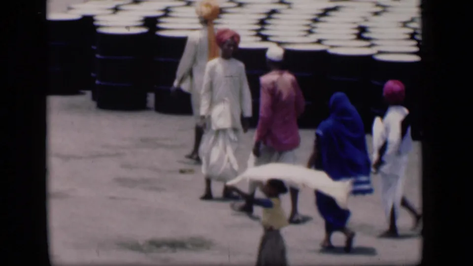 1946 Bombay street scene with pedestrians in colorful traditional garments passing rows of large dark barrels.