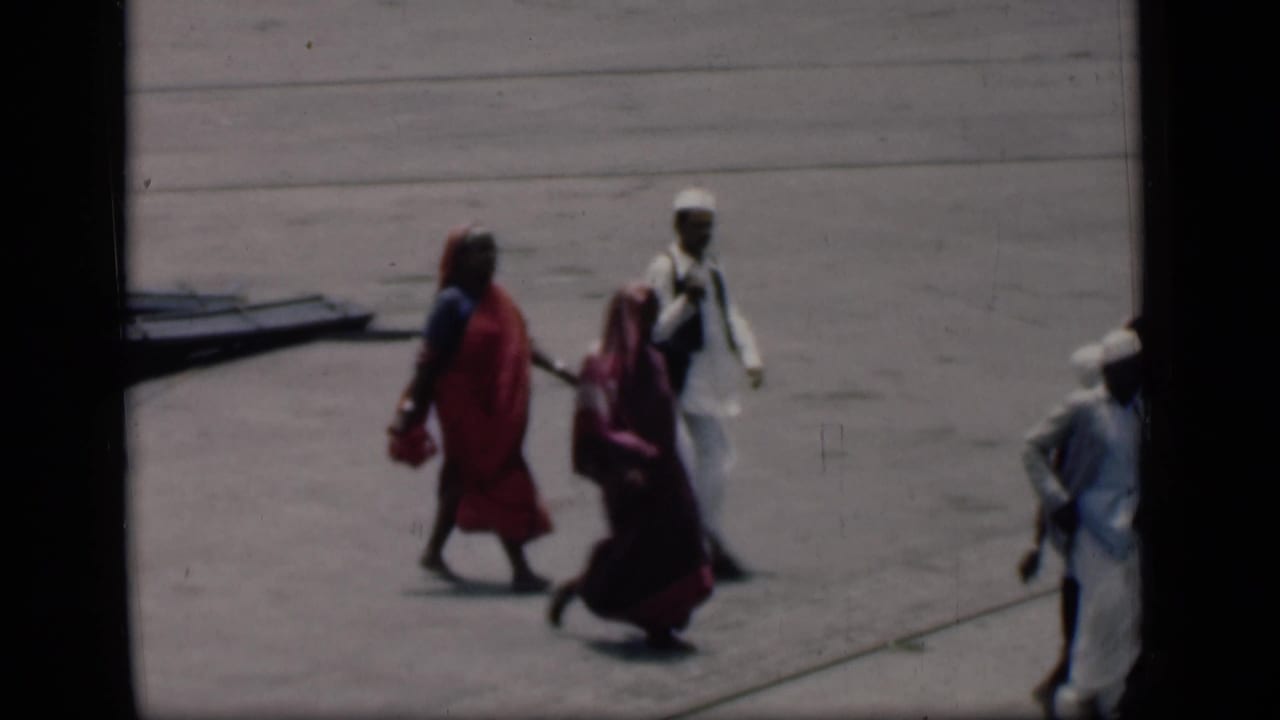 Small group walking across a sunlit tarmac, with women in sarees and men in white clothing.