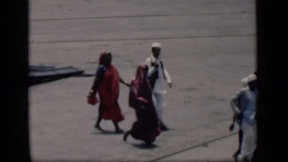 1946 Bombay home-movie frame showing people walking across an airport tarmac, including women in red and maroon sarees and men in white clothing.