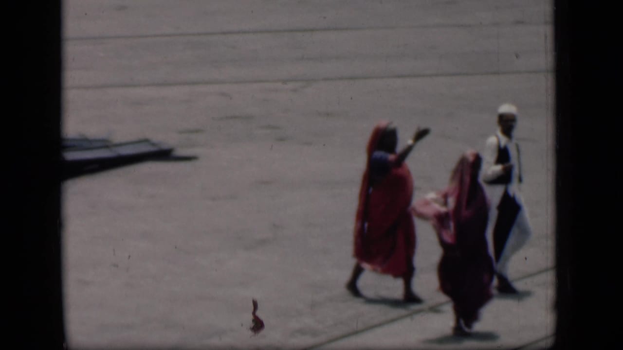 Women in sarees and a man in white walk across an open concrete tarmac.