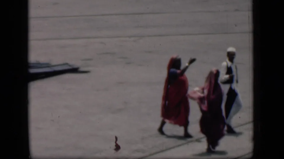 1946 Bombay airport-tarmac view with women in red and maroon sarees walking beside a man in white under bright daylight.