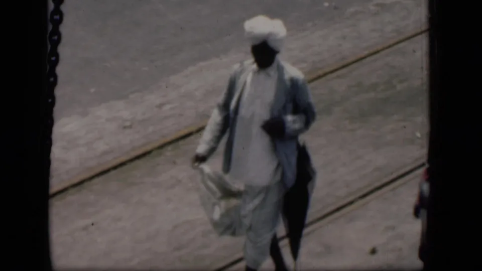 1946 Bombay frame showing a man in white traditional clothing and turban walking on the airport tarmac with a dark carried item.
