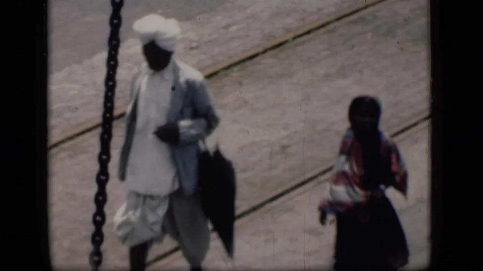 1946 Bombay airport-tarmac frame with a man in white turbaned clothing carrying a dark item and a woman in patterned dress at right.