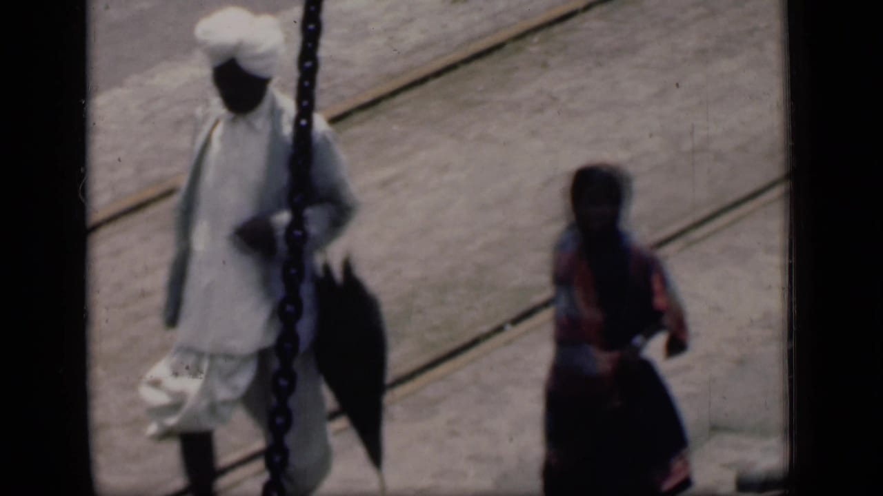 1946 Bombay dock view with a turbaned man carrying a bag and a sari-clad woman nearby on a wooden platform.