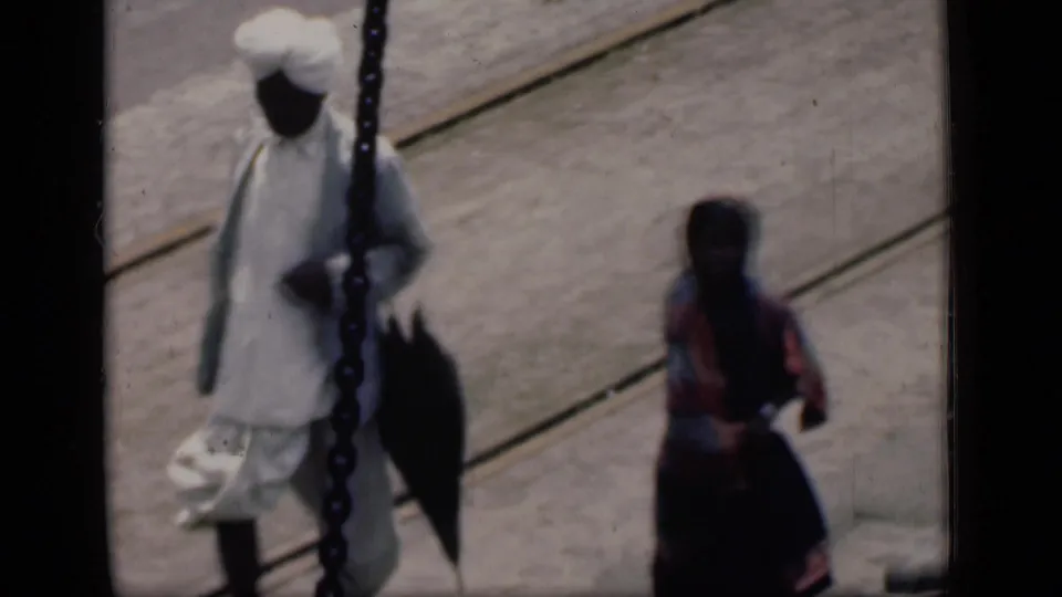 1946 Bombay dock view with a turbaned man carrying a bag and a sari-clad woman nearby on a wooden platform.