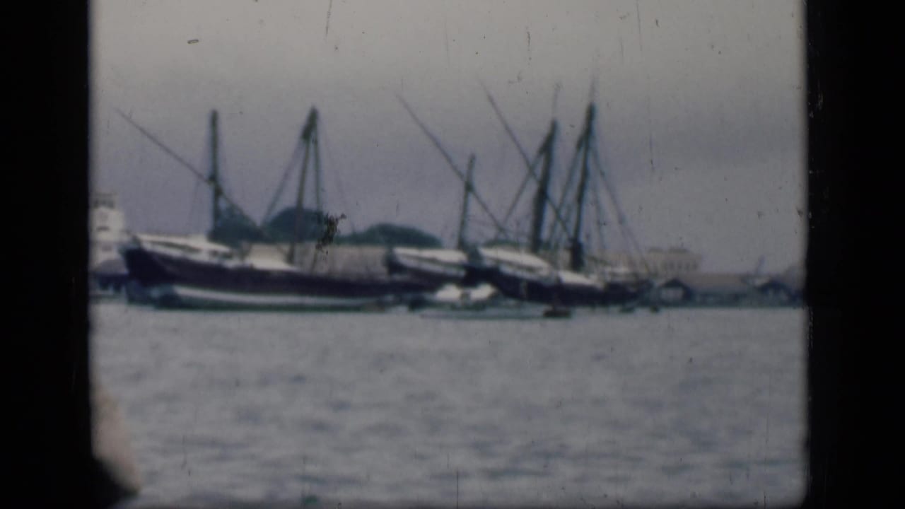 Bombay harbor view showing several moored boats with tall masts across calm water under a hazy sky.