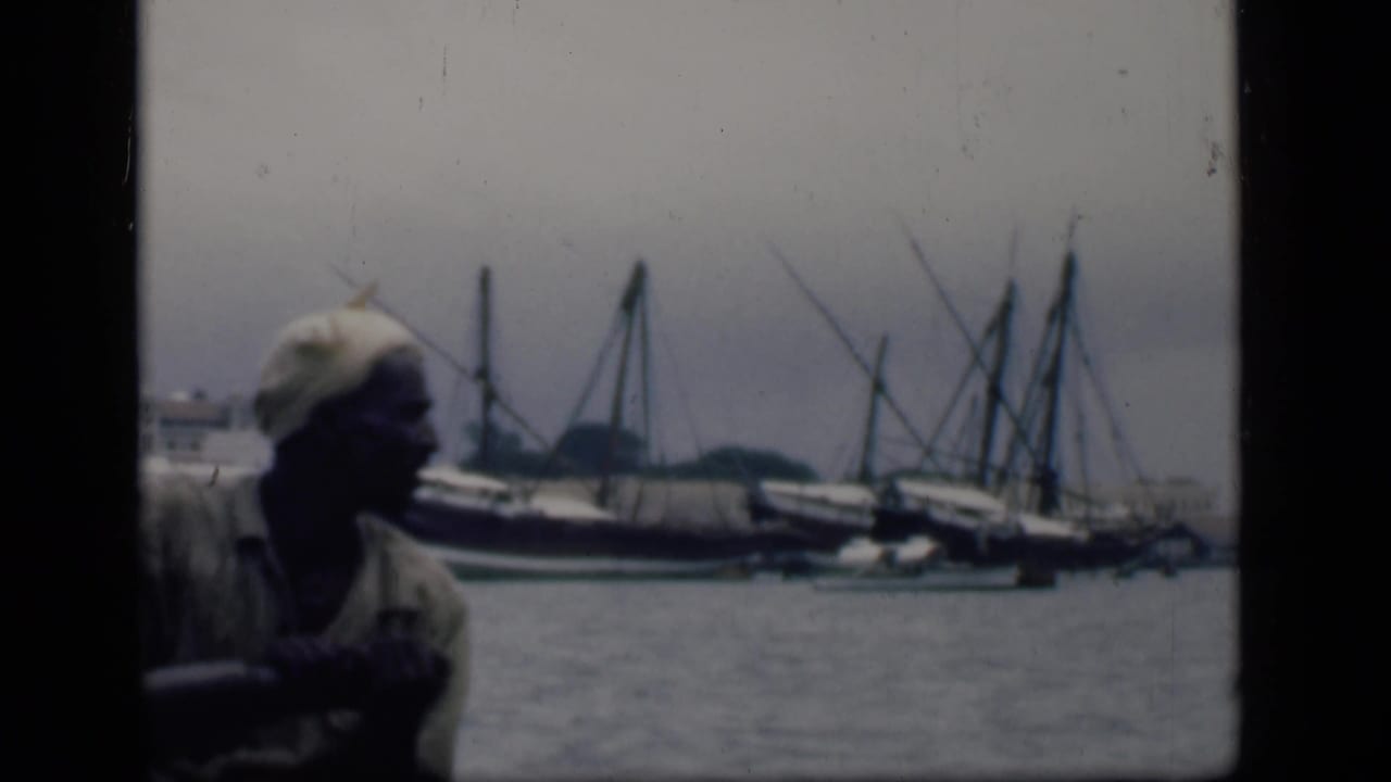 In Bombay, India, 1946, a man in a light shirt and head covering sits in a small boat with larger masted harbor boats behind him.