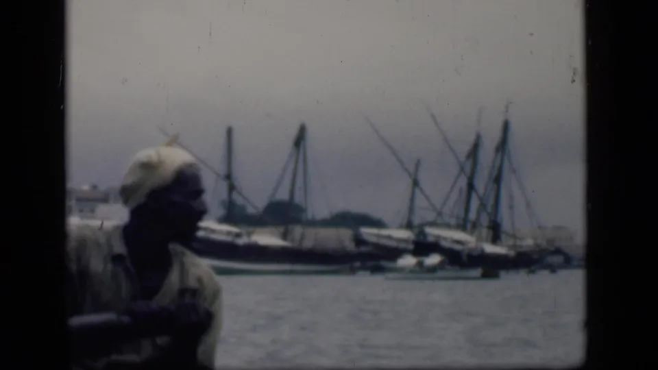 In Bombay, India, 1946, a man in a light shirt and head covering sits in a small boat with larger masted harbor boats behind him.