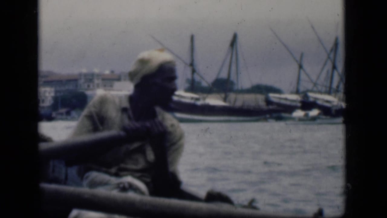 A man rows or handles an oar from a small boat in Bombay harbor, with larger traditional vessels and shoreline structures in the background.