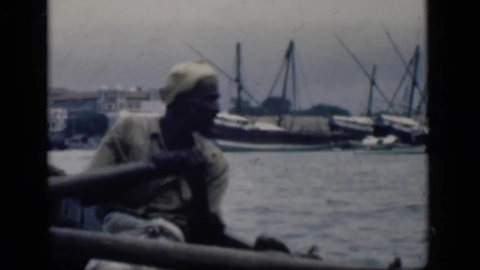 A man rows or handles an oar from a small boat in Bombay harbor, with larger traditional vessels and shoreline structures in the background.