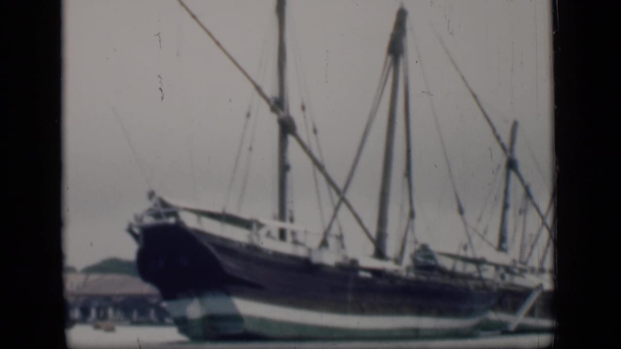 1946 Bombay frame with the bow of a large docked sailing ship, rigging, calm water, and a low shoreline building behind it.