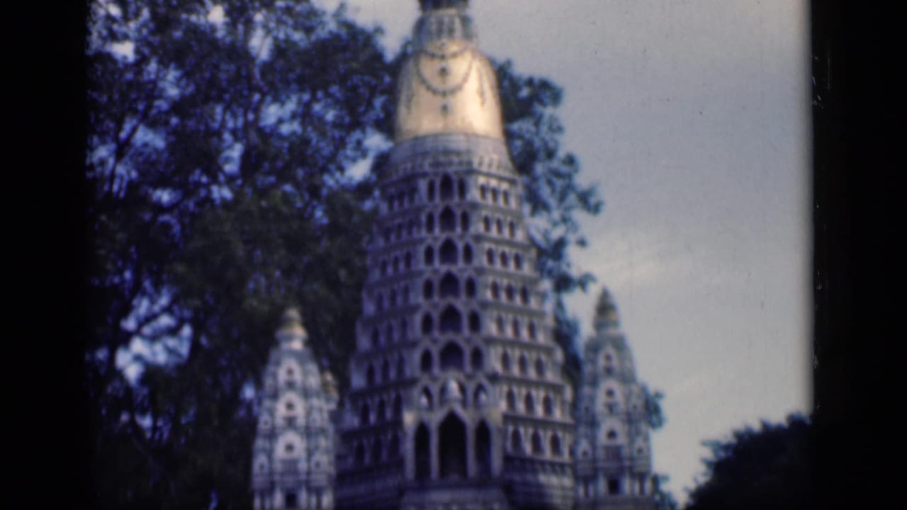 1946 Bombay 8mm frame of the temple facade, with a tall tiered tower, gold-toned upper section, trees, and pale sky.