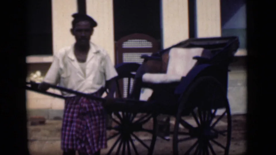 1946 Bombay home-movie frame of a man facing camera next to a rickshaw with a white cushion and large wheels.