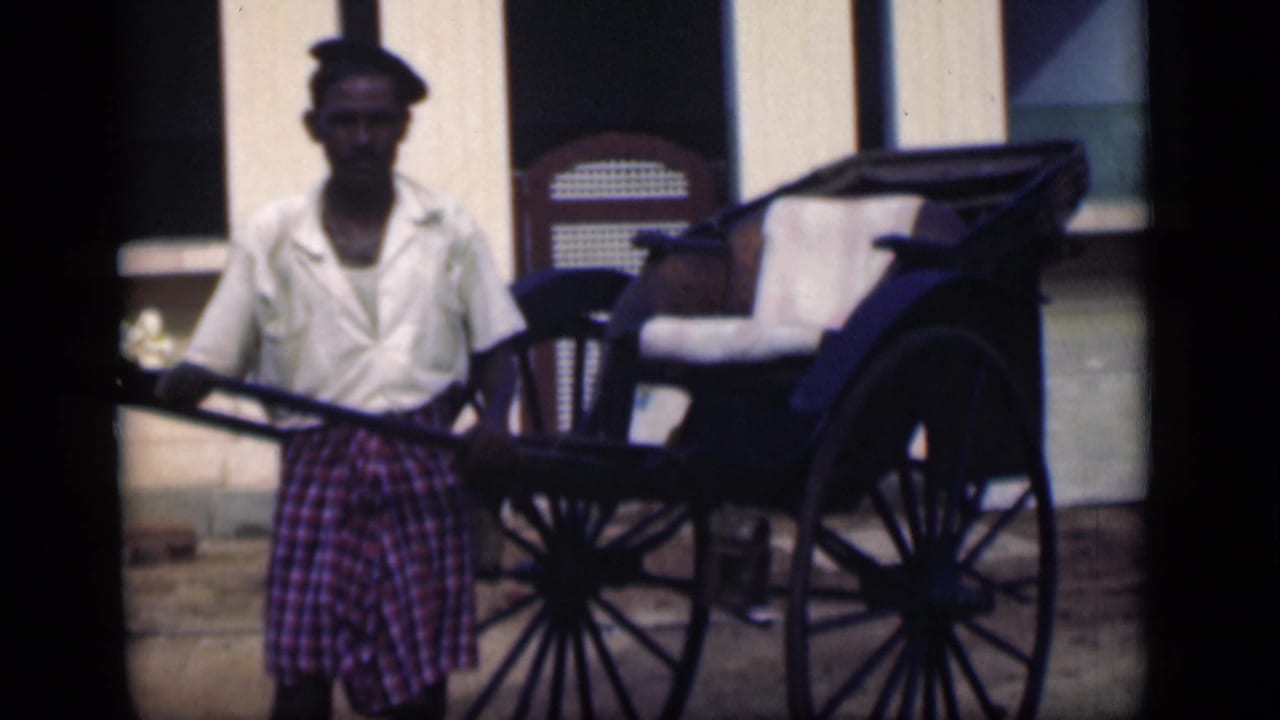 Rickshaw driver facing the camera beside a hand-pulled rickshaw and building facade.