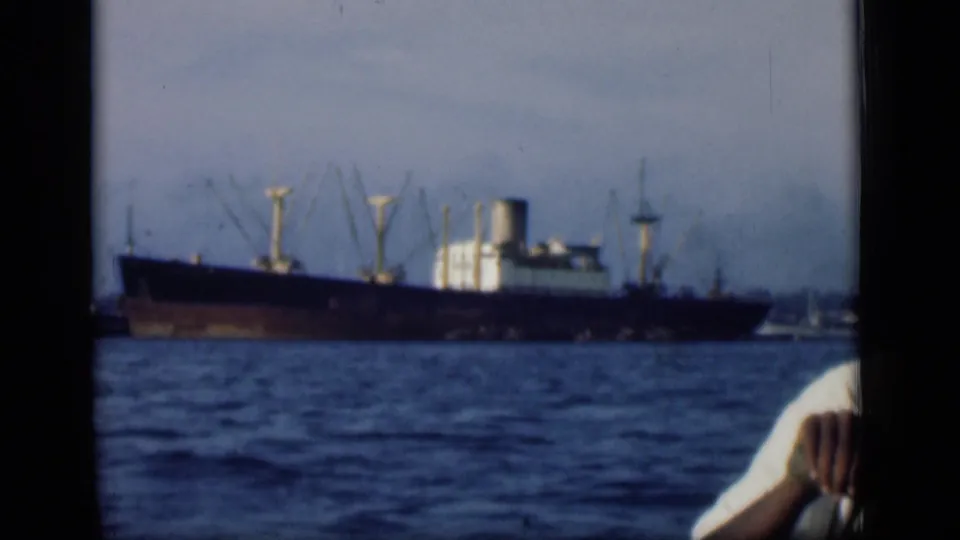 A cargo ship passes across Bombay harbor waters, seen from a boat with a foreground passenger partly visible.