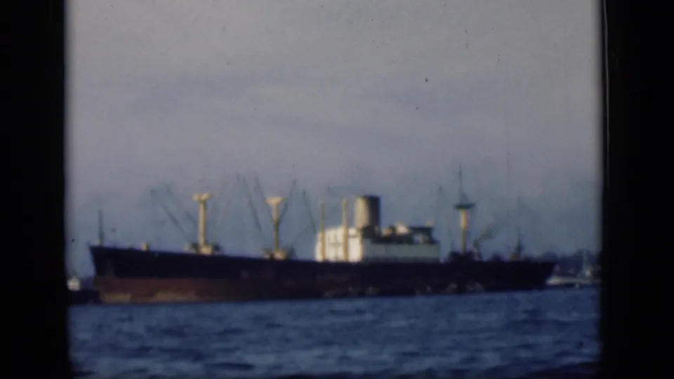 A large cargo vessel is filmed from a distance across harbor water, with port structures visible behind it.