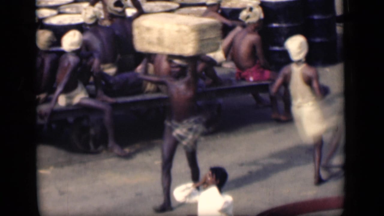 Laborers carry sacks near a wooden cart while a man in a white shirt stands in the foreground.