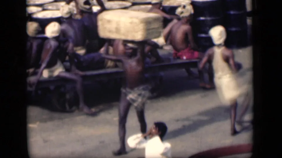 1946 Bombay home-movie frame showing men carrying head loads beside resting laborers on a wooden cart, with a white-shirted man below.