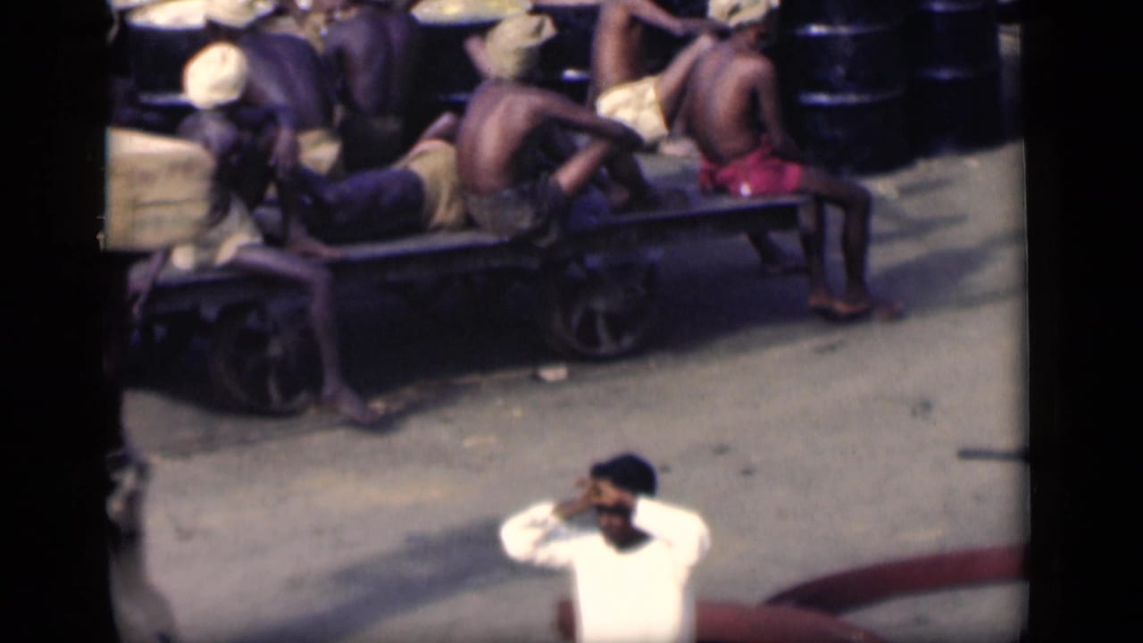 1946 Bombay frame with shirtless workers seated on a wooden cart near barrels, while a man in white raises his hand to his face.