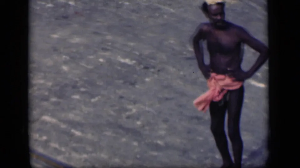 1946 Bombay home-movie frame showing a shirtless laborer standing with hands at his hips on an open paved surface.