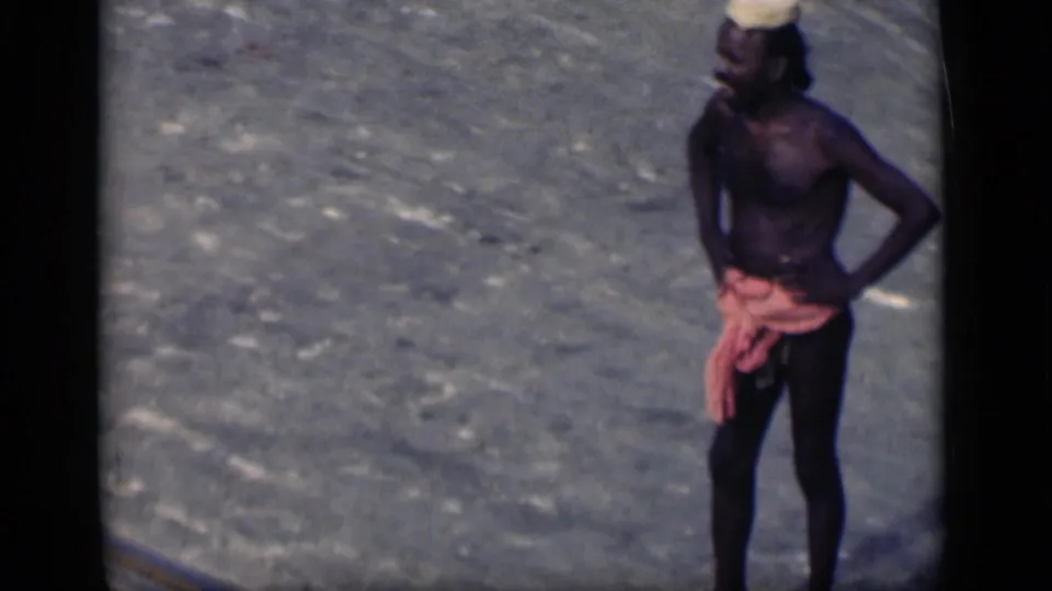 1946 Bombay frame showing a shirtless laborer in a headwrap standing on pavement, partly framed by the 8mm gate edge.