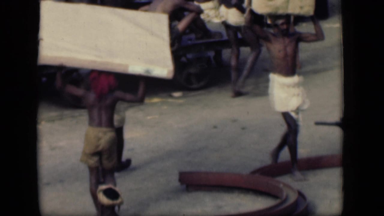 In 1946 Bombay footage, men in cloth wraps carry heavy planks or sacks on their heads near a street cart.
