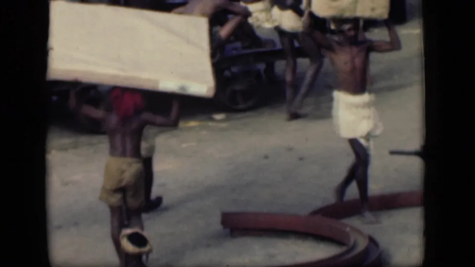 In 1946 Bombay footage, men in cloth wraps carry heavy planks or sacks on their heads near a street cart.