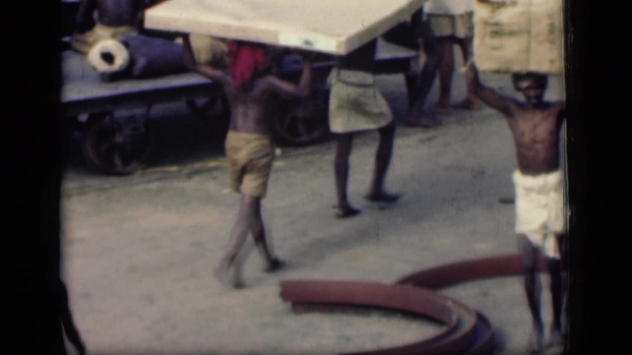 Several men walk with large head loads near a cart and street materials.