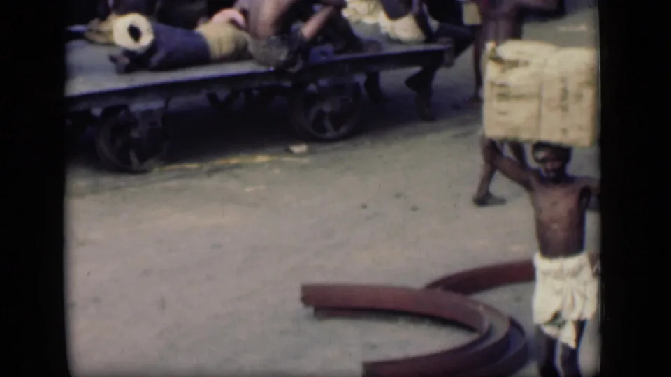 A laborer in 1946 Bombay carries a bulky head load past a wheeled cart on a dusty street.