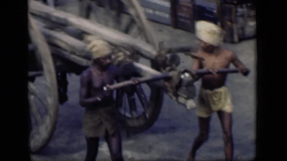 Two laborers in Bombay carry long poles or pipes past a large-wheeled cart in a close street view.