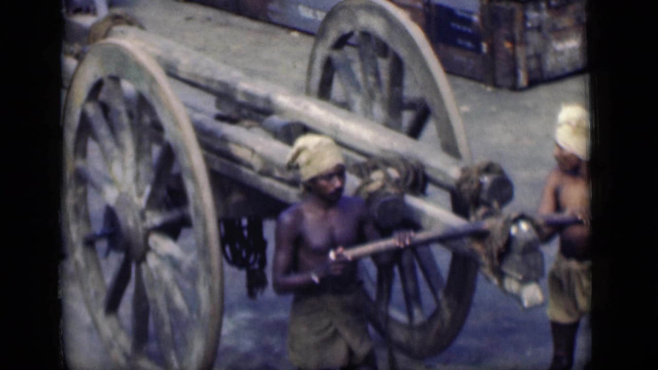 In Bombay, India, 1946, men in dhotis and headwraps work around a large wooden cart with oversized spoked wheels.