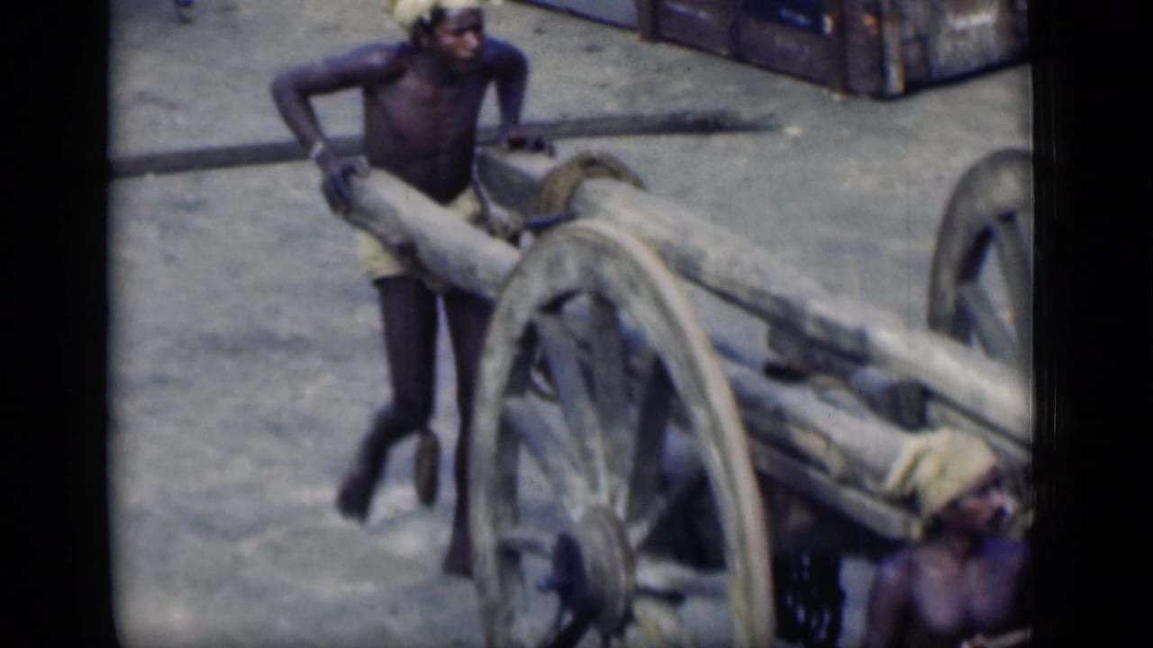 A worker in Bombay, India, 1946 appears to push or steady a heavy wooden cart as another man is partly visible nearby.