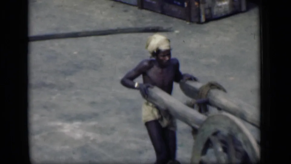 A man in a light headwrap and dhoti pushes beside a large wooden cart wheel in the 1946 Bombay street footage.