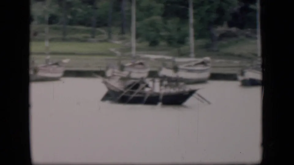 1946 Bombay 8mm frame showing small boats and oars near a grassy, tree-lined riverside bank.