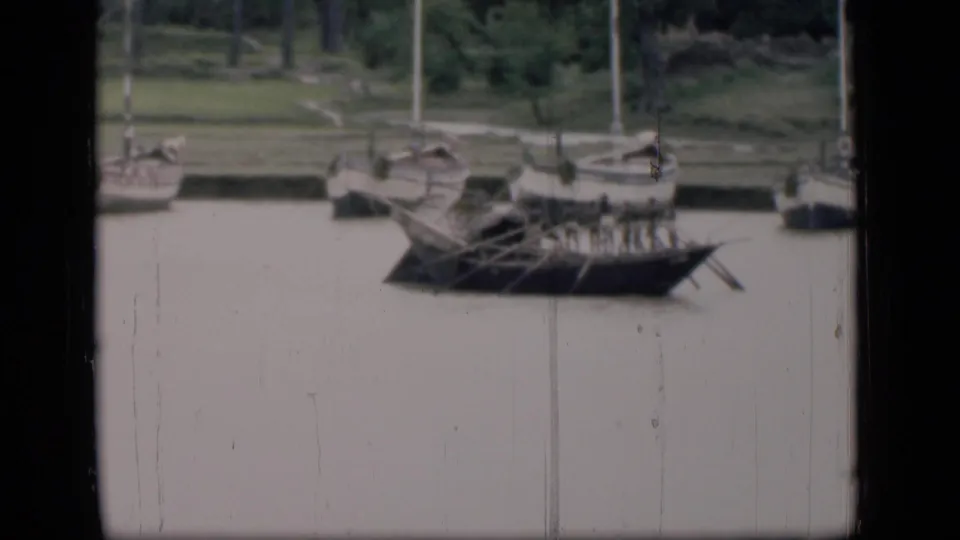 Soft 8mm view of boats along a Bombay riverside, with oars projecting from a dark wooden craft.
