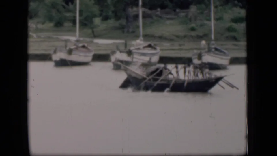 Muted 8mm frame from Bombay showing a wooden oared boat beside other small craft near a green bank.