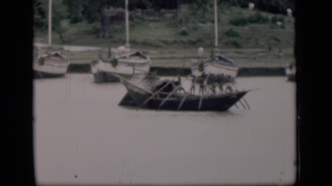 Oarsmen row a long wooden boat past moored sailboats on a river.
