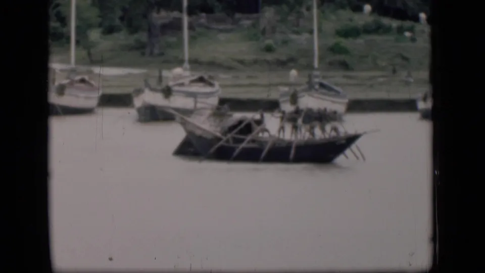 1946 Bombay home-movie frame showing several oarsmen in a long wooden boat near moored sailboats and a grassy riverbank.
