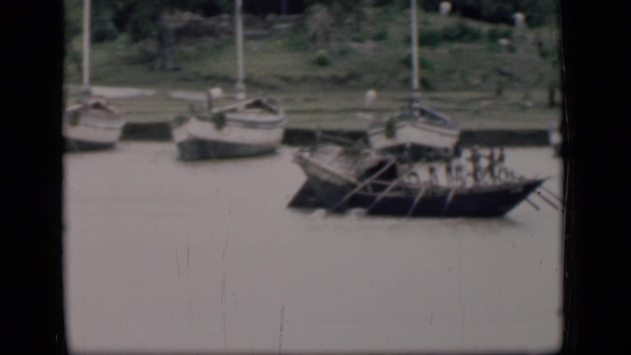A rowed wooden boat moves along the river with sailboats behind it.